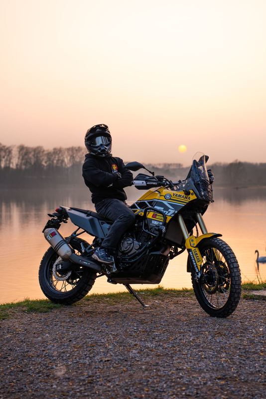 Pilote de profil sur une Yamaha Ténéré 700 en livrée rallye, au bord d'un plan d'eau au coucher de soleil